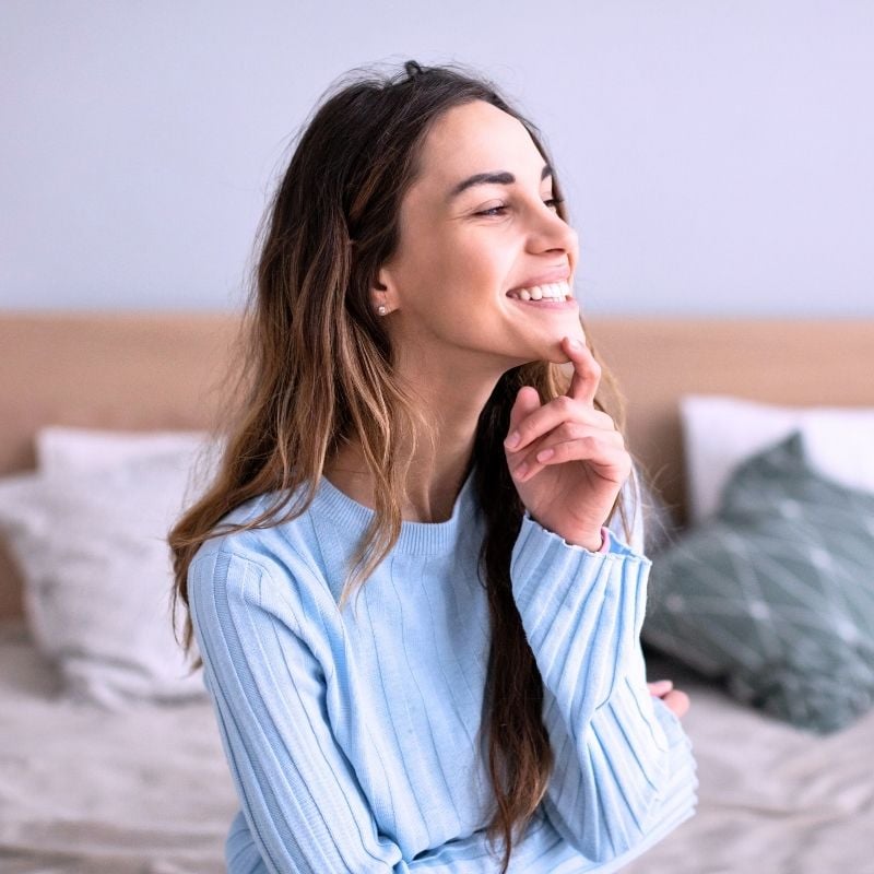 young woman sitting on bed looking out window hand to chin smiling