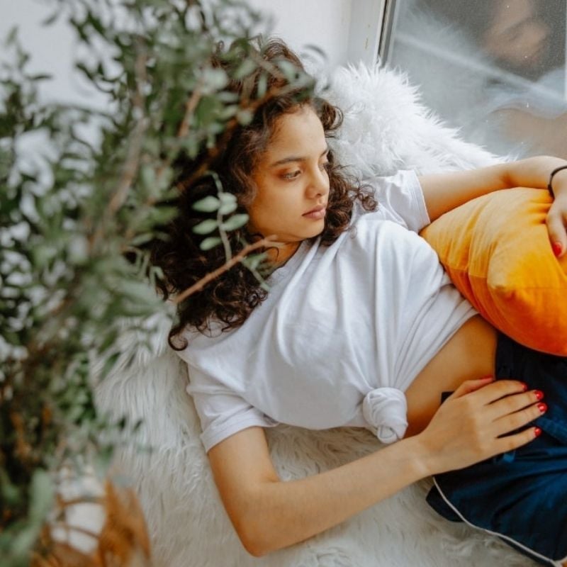 young woman lying next to window gazing out holding orange cushion over hip with one hand