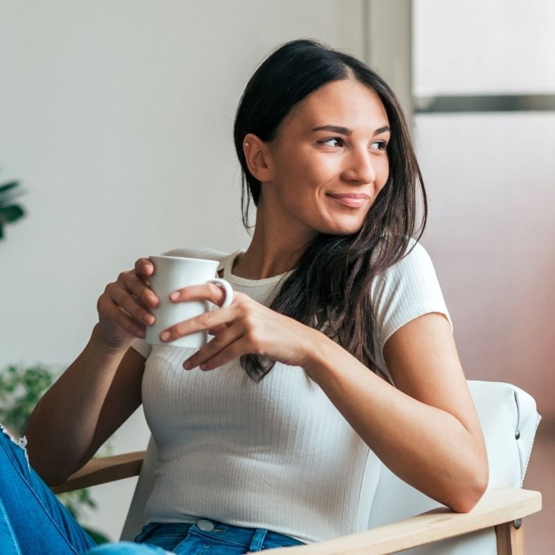 middle aged woman sitting in chair inside gazing out window holding cup tea