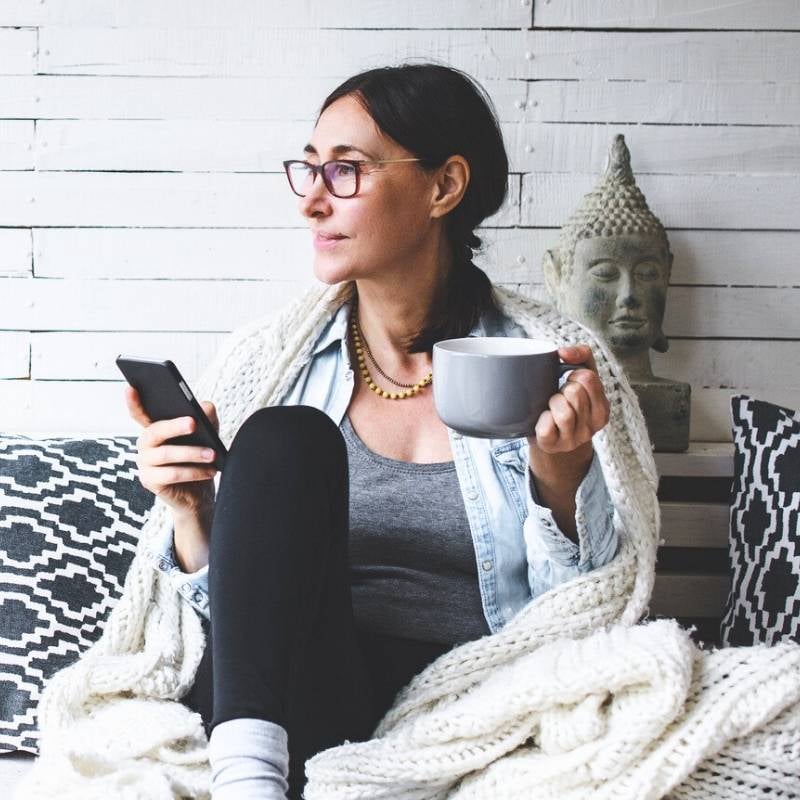 atrial-fibrillation-functional-medicine-hero woman sitting on bed with woollen blanket draped around shoulders holding mug mobile phone