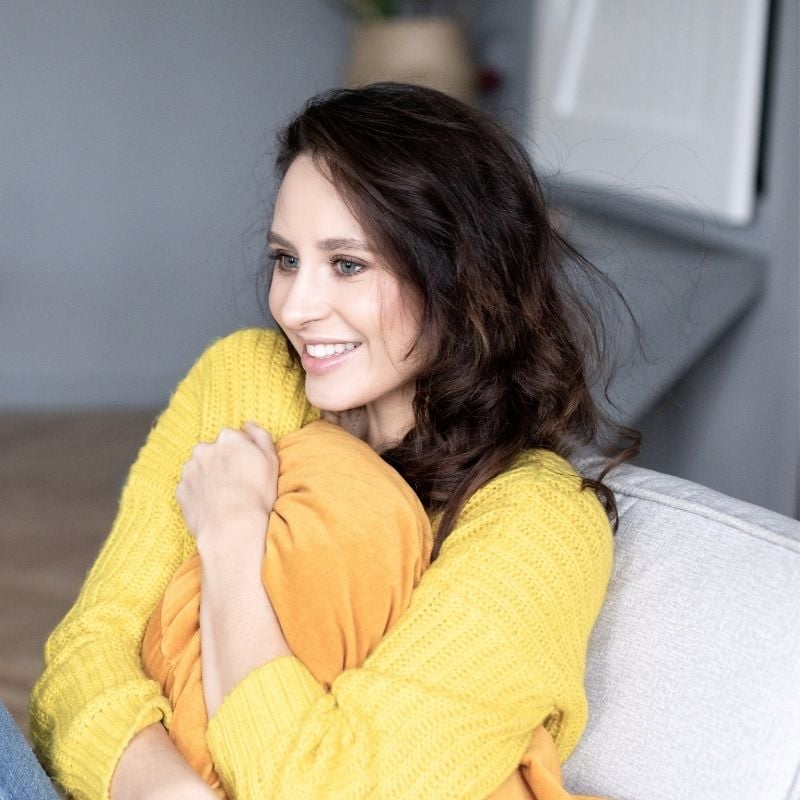 bloating-functional-medicine-hero young woman sitting on couch clutching cushion to stomach smiling
