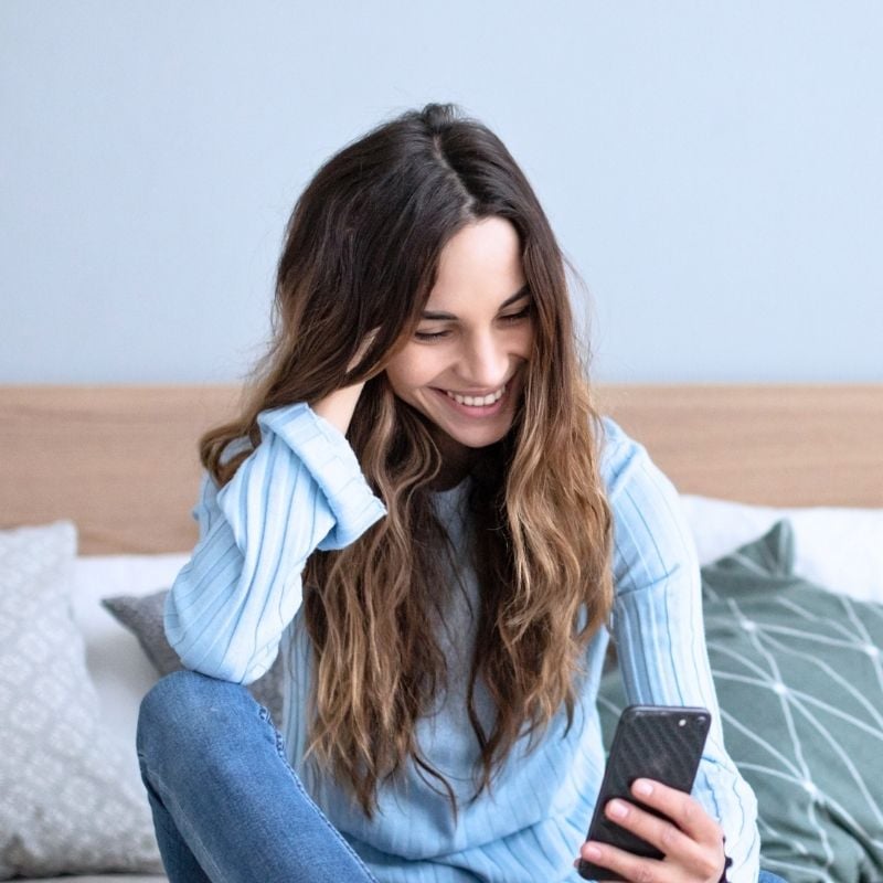 bloating-functional-medicine-root-cause young woman sitting on bed scrolling through mobile phone smiling