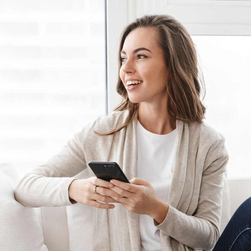 crohns-disease-sinusitis-case-study woman sitting on sofa holding mobile phone smiling looking out the window