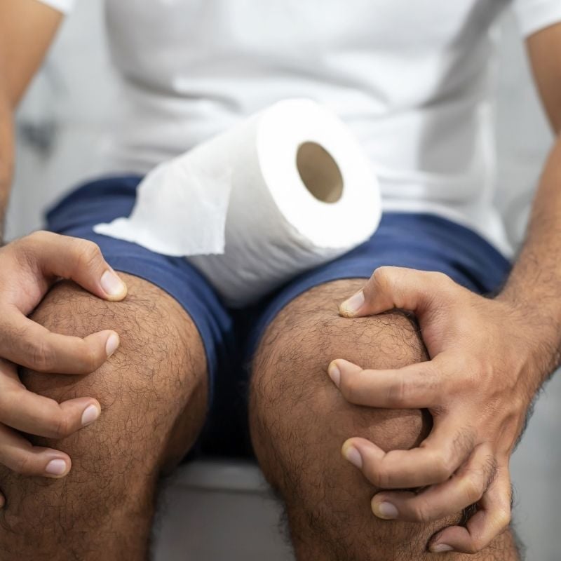 middle aged man sitting on toilet with toilet roll gripping fingers tightly around knees