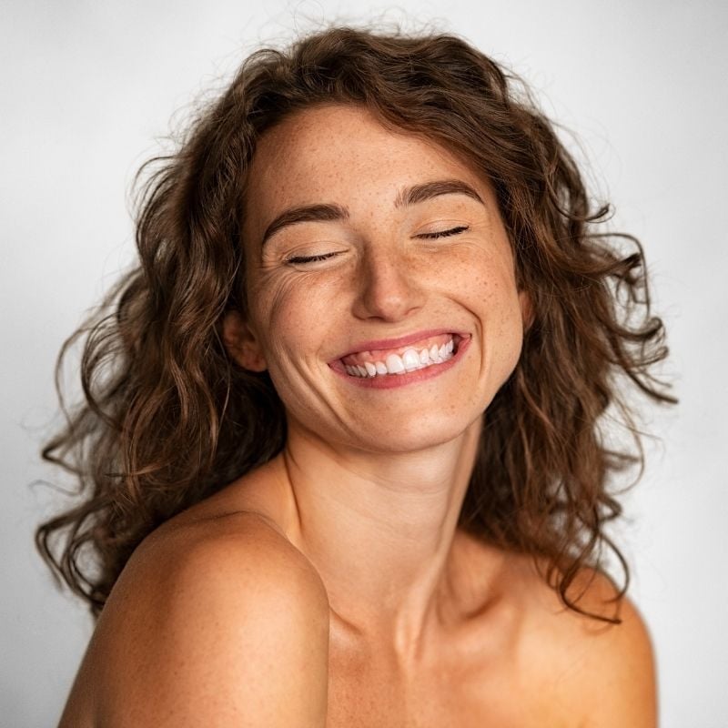 eczema-functional-medicine-root-cause woman brown wavey hair big smile eyes closed bare clear skin visible from shoulders up