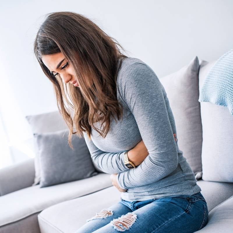 gastroparesis-functional-medicine-condition middle aged woman with food sensitivity sitting on couch arms wrapped around painful stomach