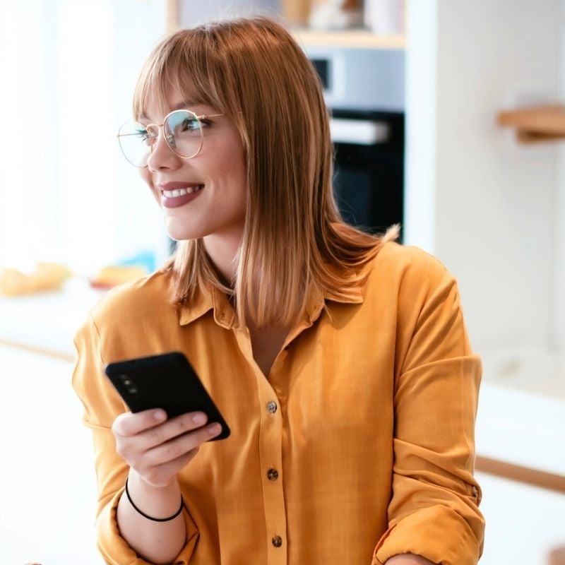 hashimotos-thyroiditis-functional-medicine-hero young woman holding mobile phone in one hand glancing out window smiling