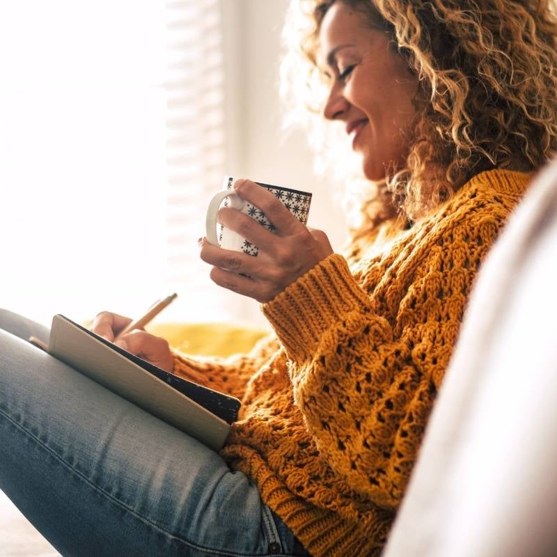 hashimotos-thyroiditis-functional-medicine-root-cause middle aged woman on couch wearing cosy jumper holding cup of tea making notes in journal