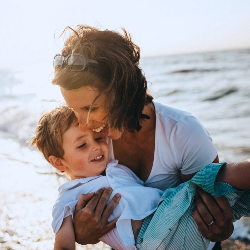 happy middle aged woman cradling young boy smiling in her arms at the beach