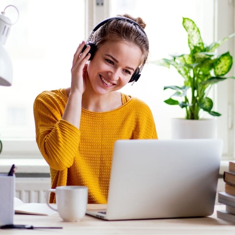 young woman sitting at table wearing headphones smiling looking at laptop