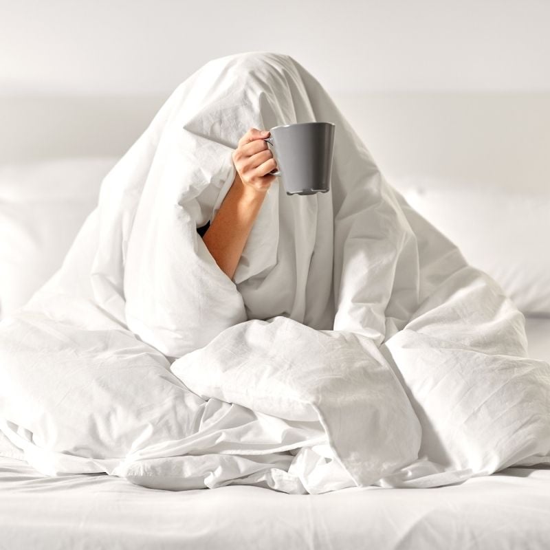 person sitting on bed with doona over head holding cup coffee