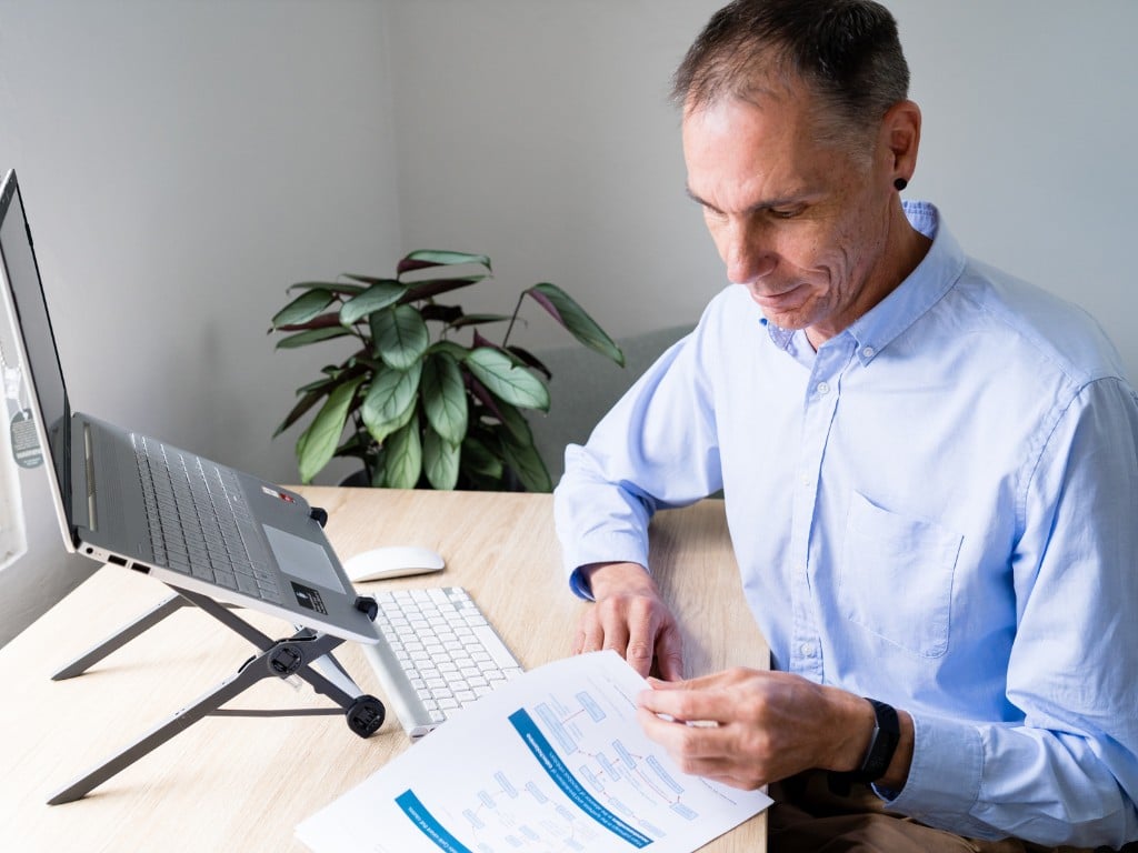 male functional medicine practitioner sitting at desk laptop open looking through print out of patient results