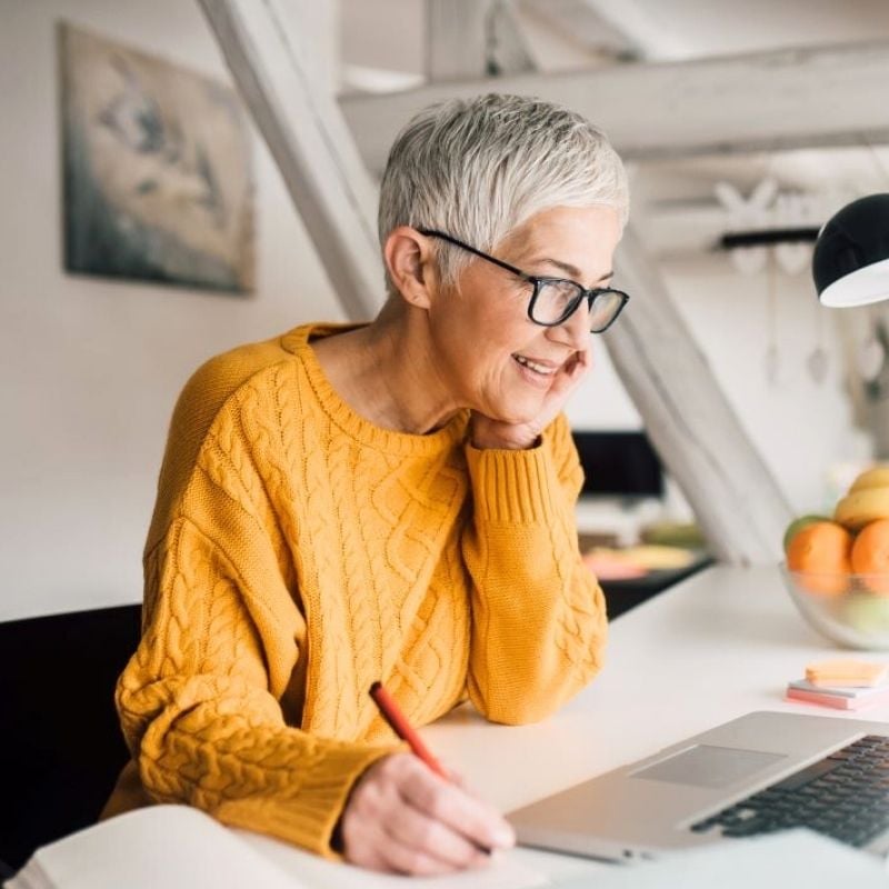 menopause-functional-medicine-hero mature woman smiling wearing glasses looking at laptop making notes in notebook with pen