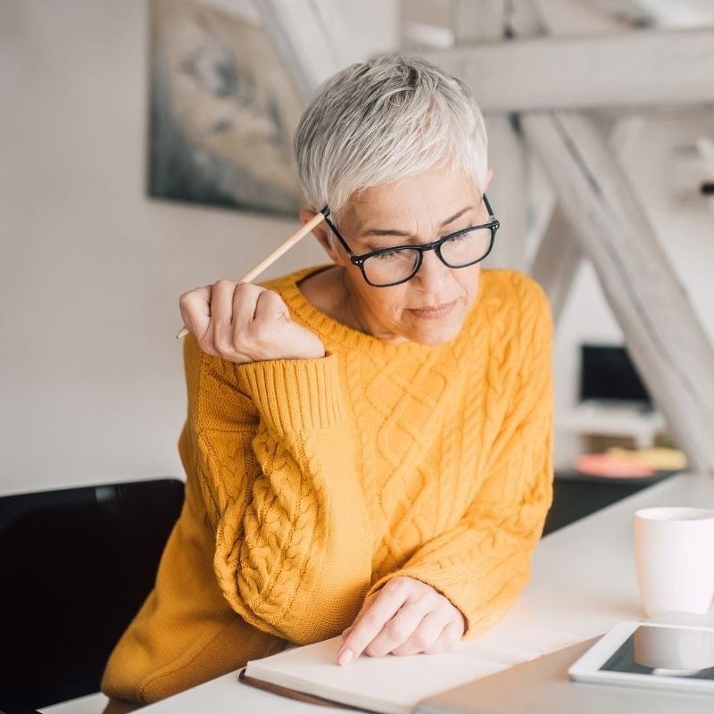 menopause-functional-medicine-root-cause mature woman sitting at desk with contemplative expression resting pencil on hairline