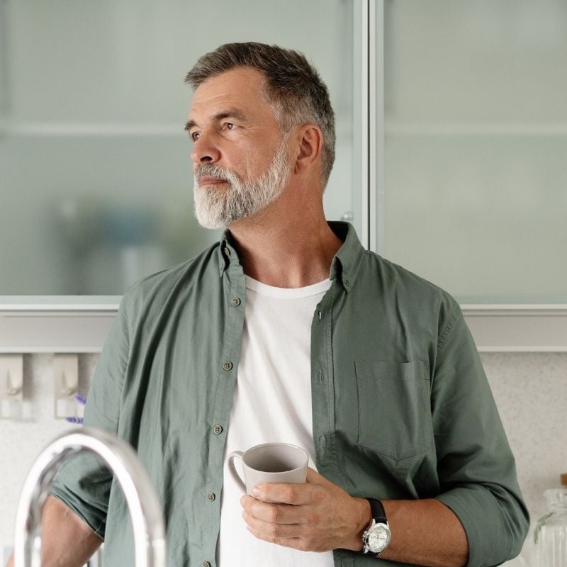 parasites-functional-medicine-condition older man standing in kitchen holding cup off with contemplative expression
