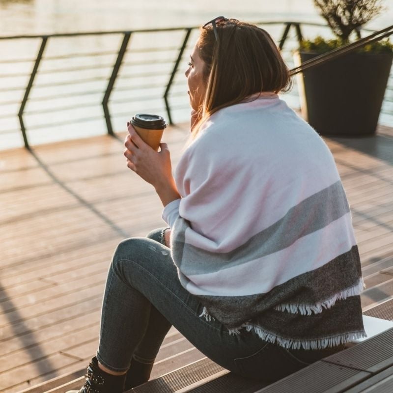 young woman sitting on decking shawl draped over shoulders watching sunrise with take away coffee
