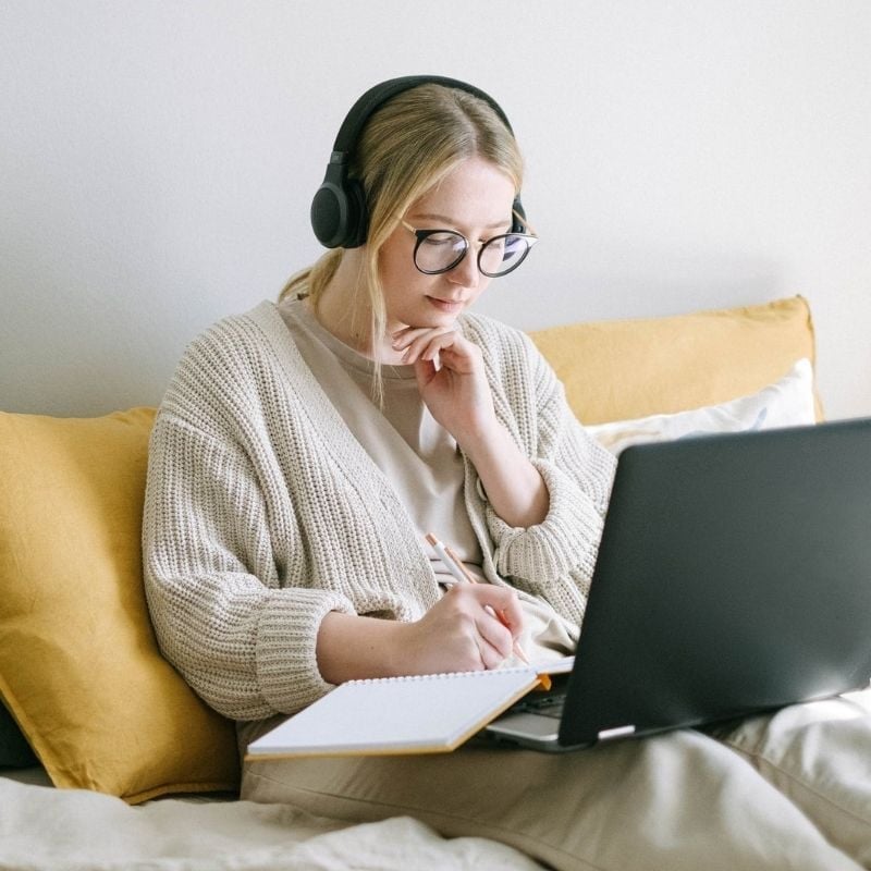 young woman sitting on couch wearing headphones looking at laptop notebook on lap