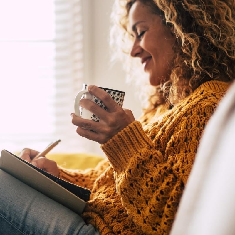 middle aged woman on couch wearing cosy jumper holding cup of tea making notes in journal