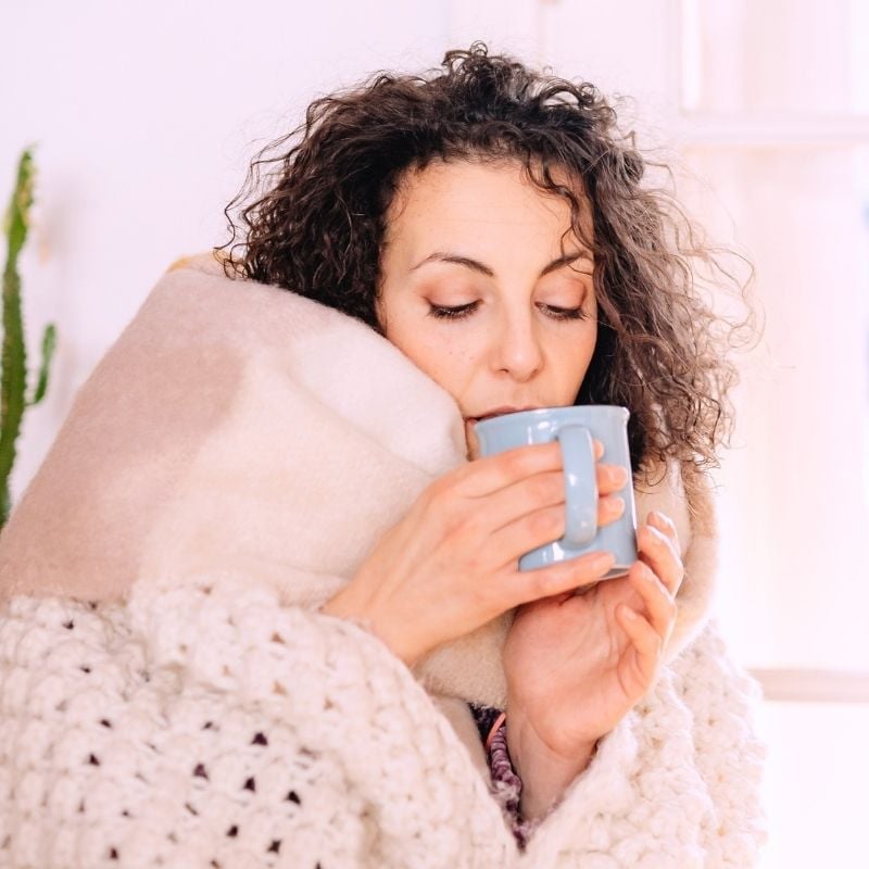middle aged woman curly hair wrapped in cosy blanket sipping sup of tea