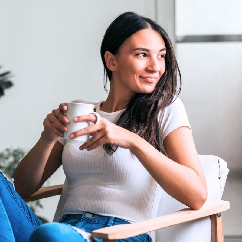 young healthy woman legs crossed on arm chair holding cup of tea smiling looking sideways out window