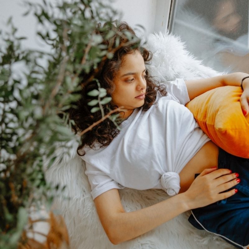 pms-functional-medicine-condition young woman lying next to window gazing out holding orange cushion over hip with one hand