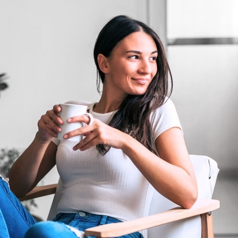 pms-functional-medicine-hero middle aged woman sitting on chair inside head turned sideways gazing out window holding cup of tea