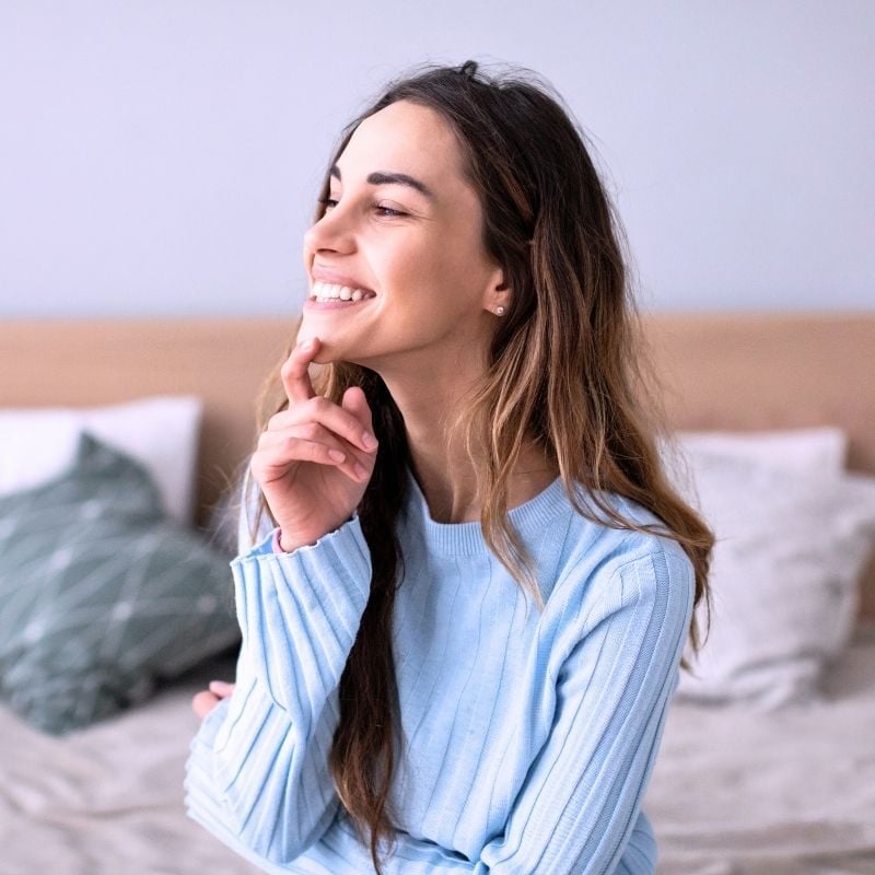 recurrent-uti-functional-medicine-hero middle aged vibrant looking woman in bright bedroom sitting on edge of bed smiling