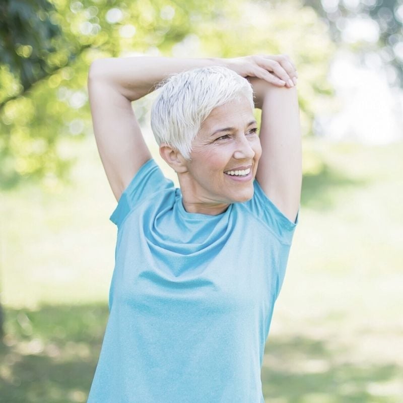 rheumatoid-arthritis-functional-medicine-hero fit older woman standing in park doing arm stretches in the sun