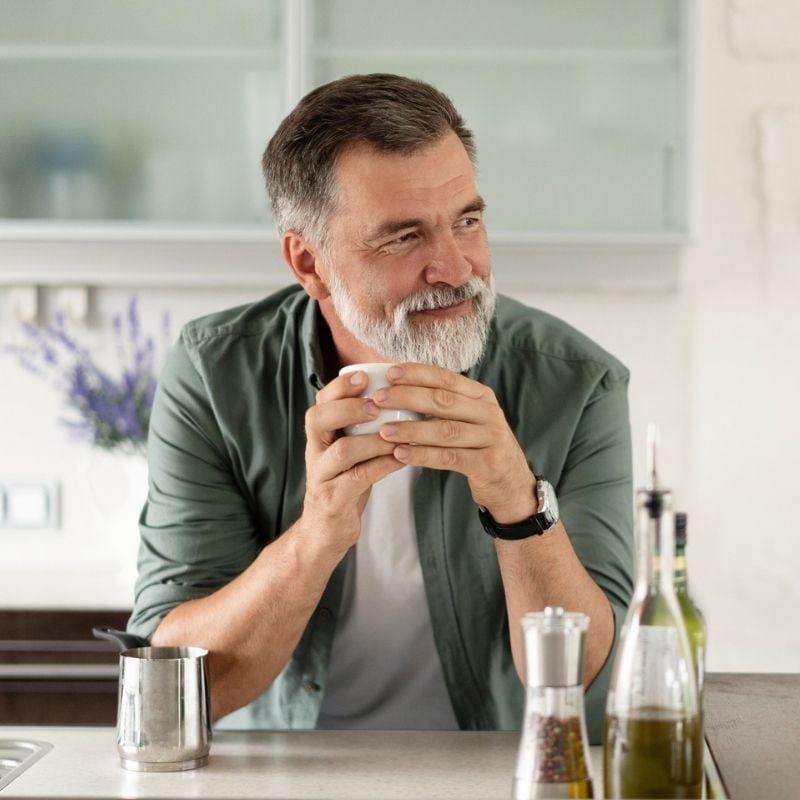 rheumatoid-arthritis-functional-medicine-root-cause older man elbows rested on kitchen bench holding cup of coffee in two hands smiling