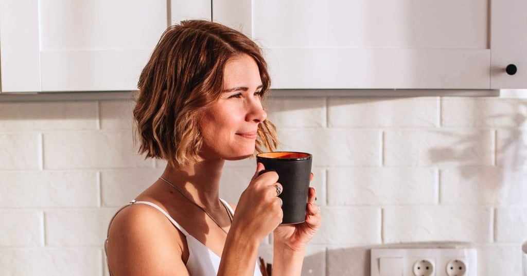 softening-the-season woman standing in kitchen holding mug up to her face sun streaming in