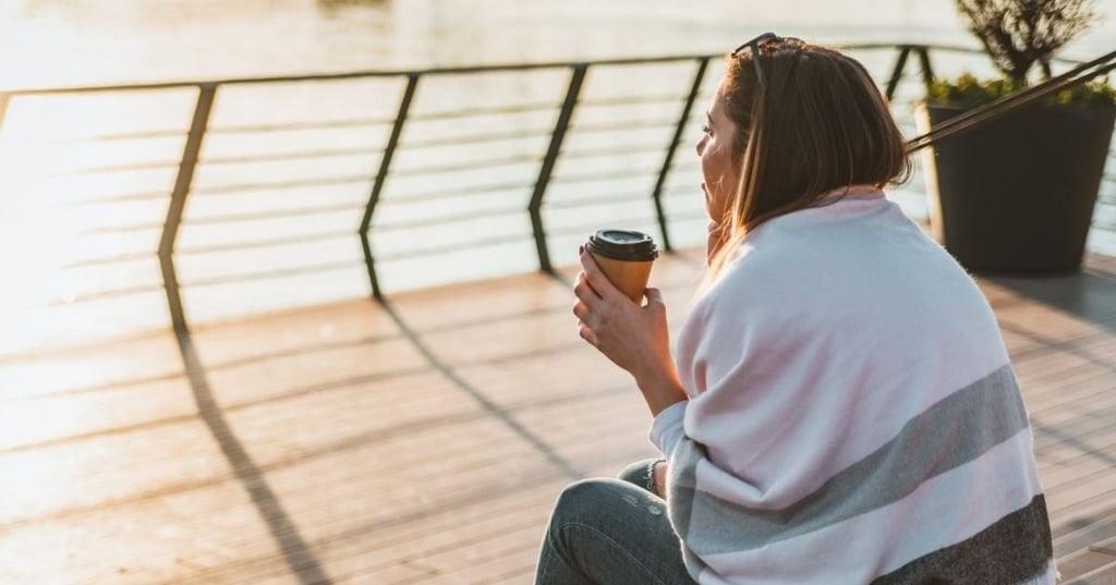 what-is-pmos young woman sitting on decking shawl draped over shoulders watching sunrise with take away coffee