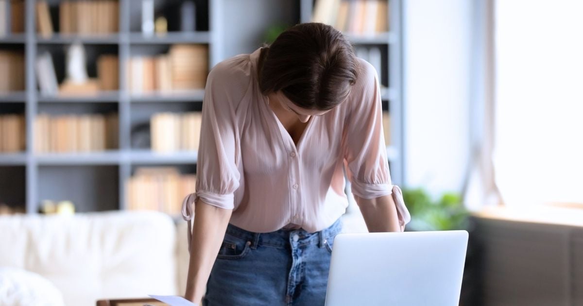 middle aged woman burnt out standing leaning over desk with head hanging forward