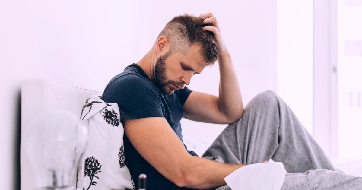 middle aged man sitting on bed head propped up with left hand looking concerned