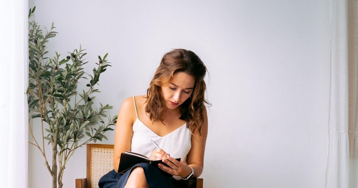 middle aged woman sitting in brightly lit room writing goals in notebook