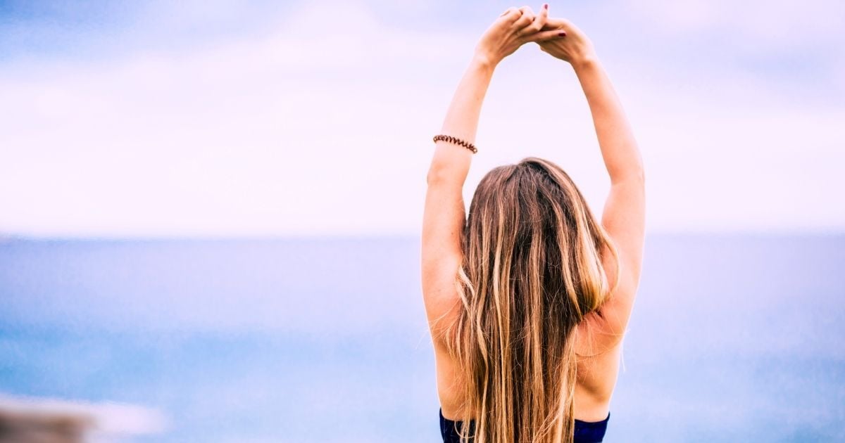 young woman in active wear standing on beach arms raised over head looking toward water