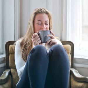 middle aged woman sitting on chair legs tucked up holding sipping cup tea both hands eyes closed
