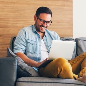 middle aged man smiling seated on couch with legs tucked up laptop on lap