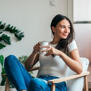 middle aged woman sitting on chair inside head turned sideways gazing out window holding cup of tea