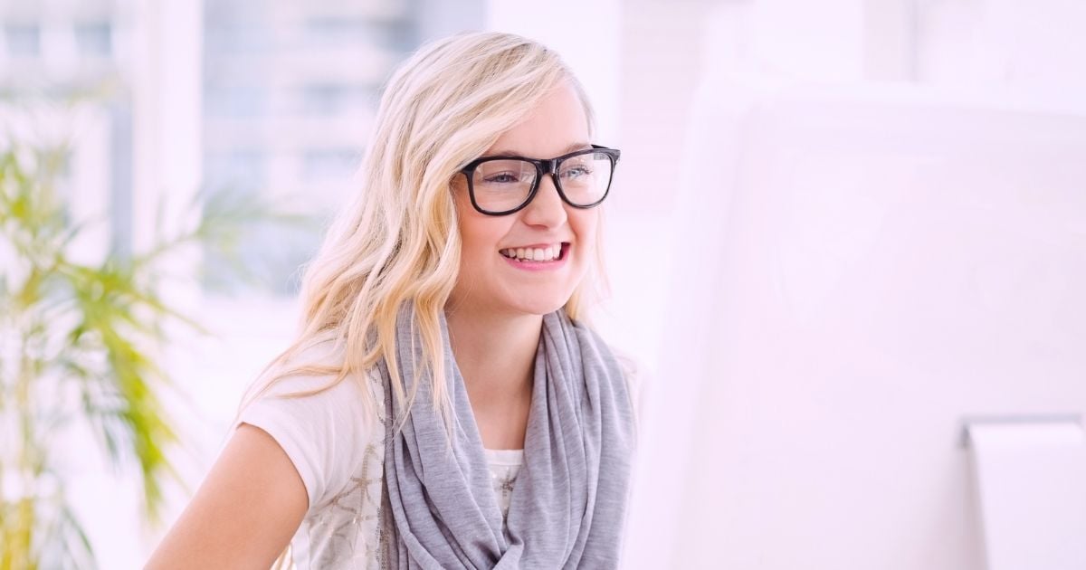young woman sitting at desk looking at computer monitor smiling right hand on mouse
