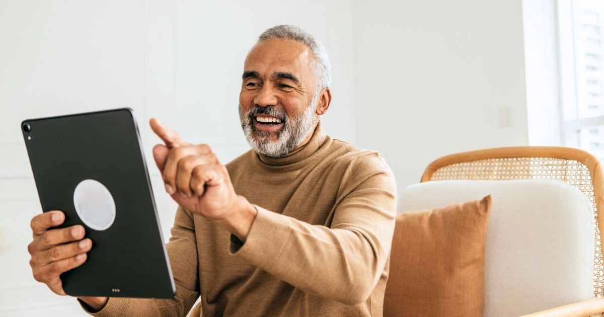 older man sitting in armchair smiling holding pointing at tablet device at arms length