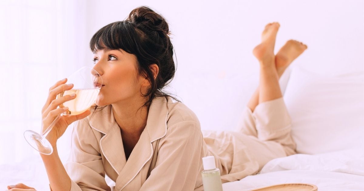 young woman lying stomach on top of bed holding glass white wine up to lips
