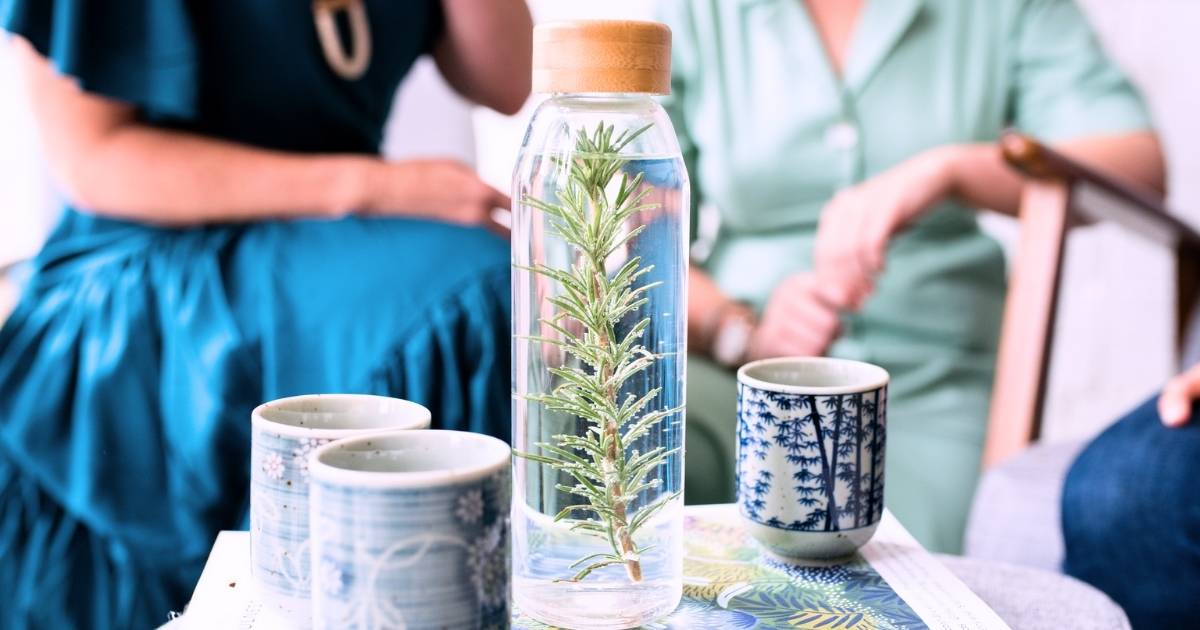 glass flask filled with water infused with sprig rosemary next to three ceramic cups