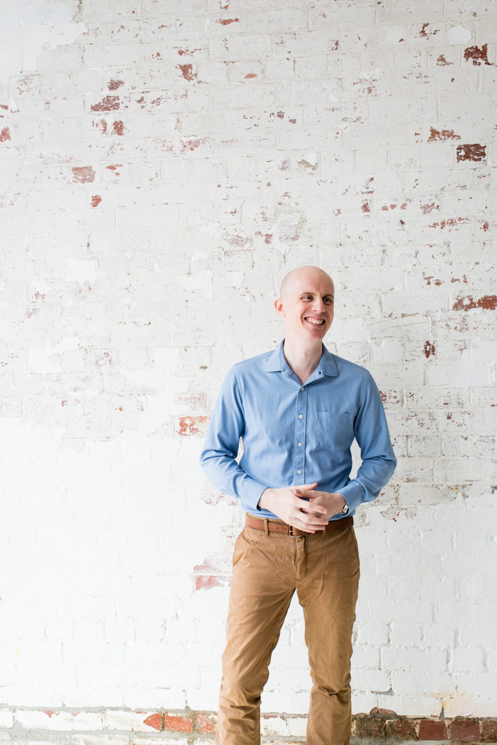 male practitioner standing in front of brick wall painted white hands clasped in front