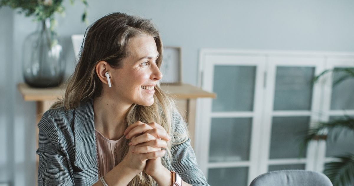 woman in midlife sitting hands clasped looking out window smiling wearing air pods