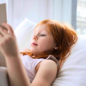 young girl lying on bed head rested on white pillow reading book