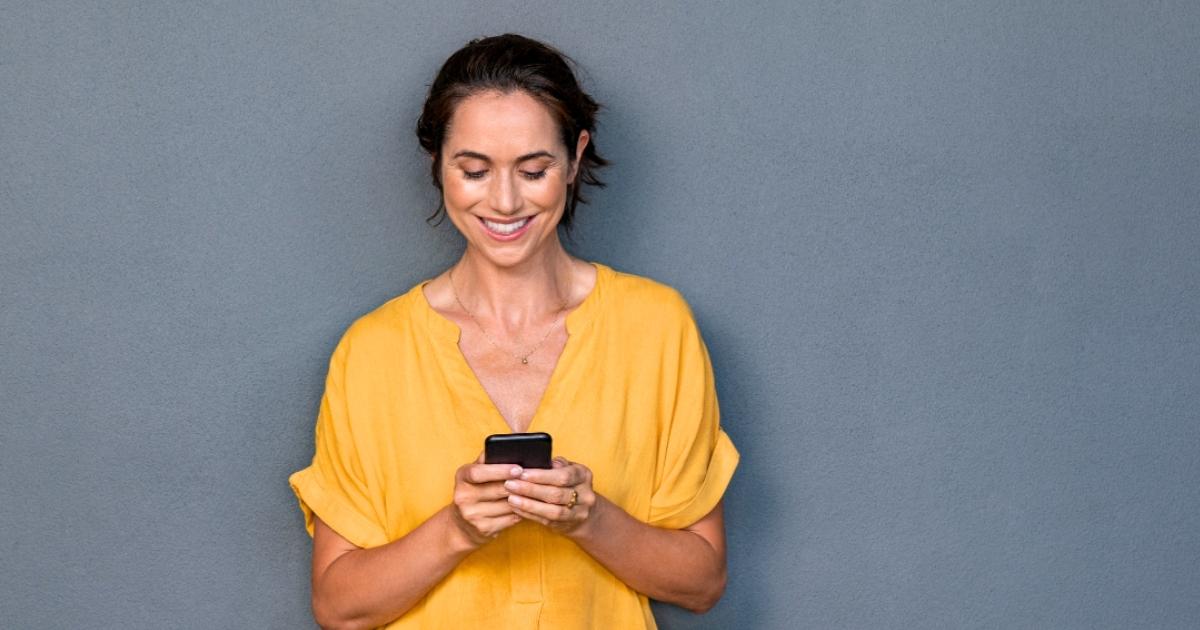 middle aged woman leaning against wall smiling scrolling through mobile phone using technology for health