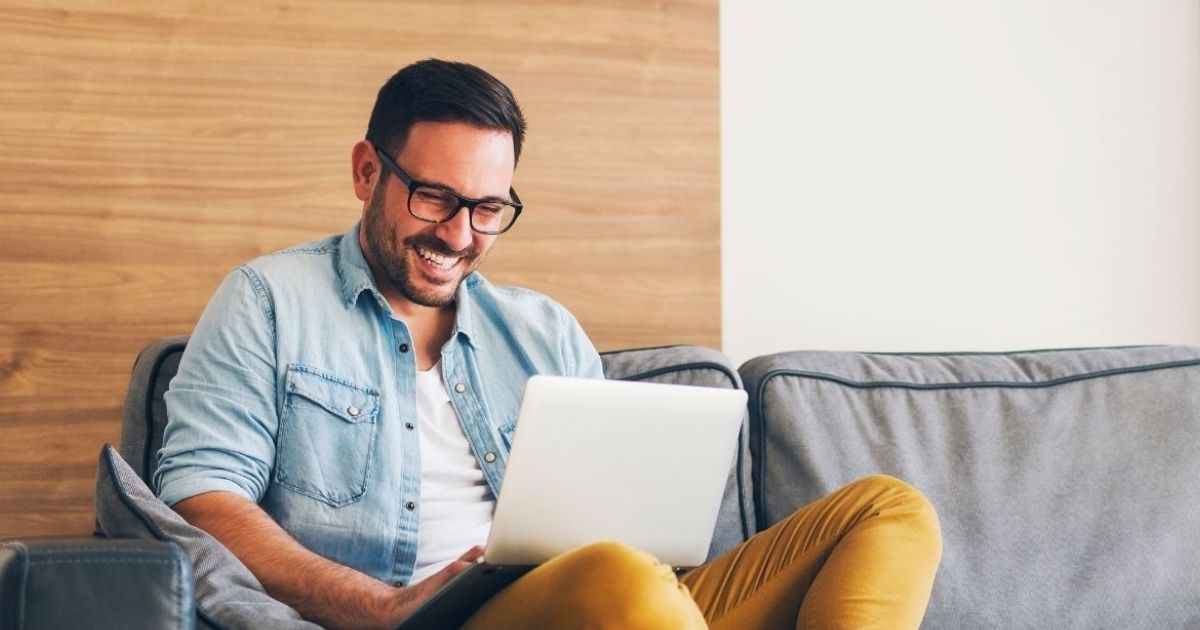 middle aged man smiling seated on couch with legs tucked up laptop on lap