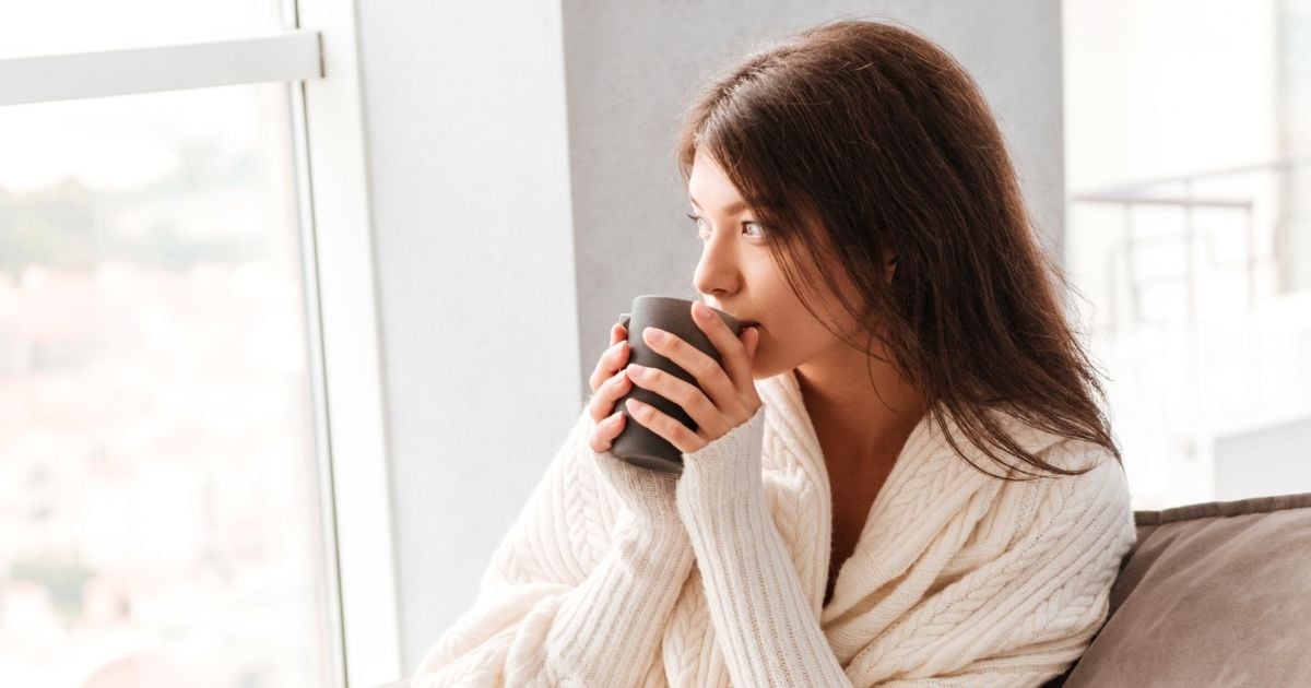 woman sitting on couch looking out window holding drink with two hands